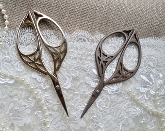 May include: Two decorative scissors with ornate leaf-shaped handles. One is gold-toned, the other is bronze. The scissors are set against a backdrop of lace and burlap, suggesting a craft or sewing theme.