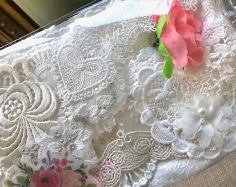May include: Close-up of a collection of white lace fabric pieces with various floral and heart designs. A pink flower with green leaves is attached to the top. The lace appears to be delicate and intricate.