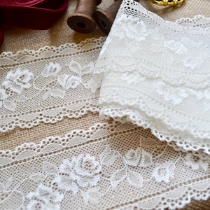 May include: Close-up of white lace trim featuring a floral rose design. The lace is layered and folded, with a scalloped edge. The background includes a burlap surface and spools of thread.