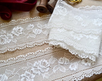 May include: Close-up of white lace trim with a floral rose design. The lace is layered and folded, with a scalloped edge. The background includes a burlap surface and spools of thread.