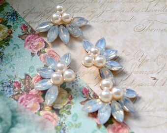 May include: Close-up of several floral-shaped embellishments. Each features opalescent blue-toned petals and a cluster of three faux pearls at the center, set on a gold-colored base. The background includes floral and script patterns.