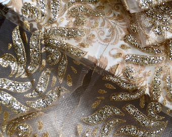 May include: Close-up of a luxurious fabric with a gold and silver glitter design. The fabric features a cream-colored base with a gold floral pattern, overlaid with a sheer layer adorned with gold glitter accents. The overall effect is elegant and ornate.