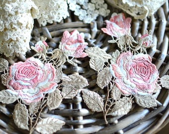 May include: Embroidered floral appliques featuring pink roses with detailed stitching and gold-toned leaves. The appliques are set against a woven background, with lace trim visible in the upper corners. These decorative appliques are ideal for craft projects.