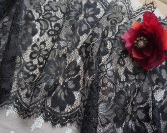 May include: A length of black lace fabric featuring a detailed floral pattern with scalloped edges. A single deep red artificial flower with a central pearl-like embellishment is pinned to the right side of the delicate, semi-sheer material.
