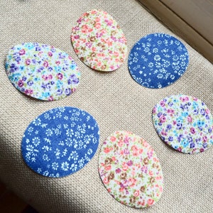 Puede incluir: Seis parches de tela en formas ovaladas y circulares, cada uno con un estampado floral. Los estampados incluyen pequeñas flores en tonos rosa, azul, morado y blanco sobre un fondo claro. Los parches están dispuestos sobre una tela beige.