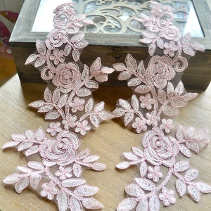 May include: Two pink floral lace appliques featuring rose and leaf patterns. These delicate, embroidered lace appliques are suitable for embellishing clothing or crafts. The appliques are displayed on a wooden surface.