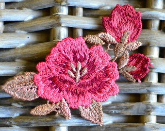 May include: Close-up of a red embroidered flower with gold-colored leaves and details. The flower is set against a woven, light brown background, creating a contrast of textures and colors. The embroidery is detailed and textured.