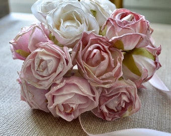 May include: A bouquet of artificial roses in shades of pink and white, with a satin ribbon. The roses have a textured, slightly glittery appearance. The arrangement is set against a neutral background.