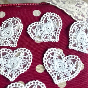 Può includere: Primo piano di applicazioni a forma di cuore in pizzo bianco con disegni floreali, su uno sfondo bordeaux a pois. I cuori di pizzo hanno un bordo smerlato. Lo sfondo è una superficie strutturata e imbottita.