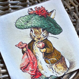 Peut inclure: Écusson carré en tissu avec une illustration détaillée d'un lapin portant un chapeau vert et une veste marron. Le lapin tient un paquet rouge et un petit fruit. L'œuvre a un style vintage de livre d'histoires.