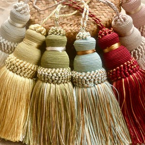 May include: A collection of decorative tassels in various colors, including gold, green, red, and pale pink. Each tassel features a bulbous top with intricate detailing and a long, flowing fringe. The tassels are arranged on a woven background.