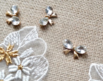 May include: Close-up of decorative embellishments. Clear, teardrop-shaped rhinestones are set in gold-colored metal bows. White lace floral appliques with gold centers are also visible, all on a textured beige fabric.