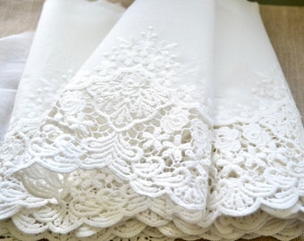 May include: Close-up of white lace fabric with intricate floral and scrollwork patterns. The lace is layered, creating depth and texture. The delicate design is visible along the edges, showcasing the craftsmanship. The fabric appears soft and lightweight.
