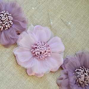 May include: Three fabric flowers in shades of purple and lavender, with a layered petal design. The center of each flower has a textured, rolled appearance. Two sewing pins with white heads are also visible, suggesting a craft or DIY project.
