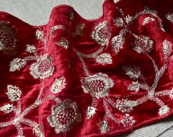 May include: Close-up of a deep red velvet fabric with intricate silver floral embroidery. The embroidery features detailed flowers and leaves, creating a luxurious and ornate design. The fabric is likely used for decorative purposes.