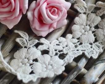 May include: Close-up of cream-colored lace trim with a floral design, alongside two pink roses. The lace features detailed embroidered flowers and leaves. The items are arranged on a woven, light brown surface, creating a delicate and vintage aesthetic.