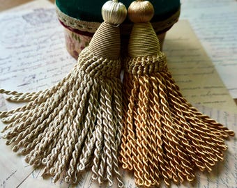May include: Two decorative tassels featuring twisted cord fringe, one in a pale champagne gold and the other in a deeper metallic gold. Each tassel has a rounded, wrapped top section and a braided neck, finished with a matching cord loop at the top.
