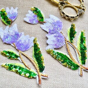 May include: Embroidered floral appliques featuring lavender sequined flower petals and green sequined leaves. Gold thread outlines the stems and leaves. A pair of gold scissors and purple velvet ribbon are also visible.