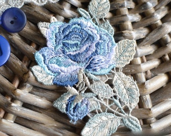 May include: Close-up of a blue embroidered rose applique with detailed stitching and delicate leaves. The applique is set against a woven background, with blue buttons and lace trim visible. A beautiful floral embellishment.