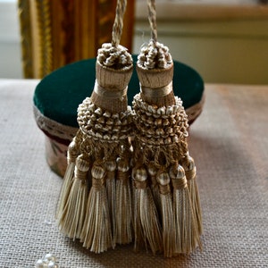 May include: Two decorative gold tassels with intricate detailing. Each tassel features a knotted top, a beaded midsection, and multiple strands of flowing threads. The tassels are displayed against a neutral background, suggesting a focus on home decor or craft supplies.