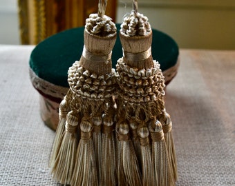 May include: Two decorative gold tassels with intricate detailing. Each tassel features a knotted top, a beaded midsection, and multiple strands of flowing threads. The tassels are displayed against a neutral background, suggesting a focus on home decor or craft supplies.