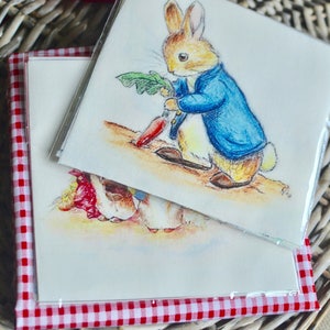Puede incluir: Dos tarjetas cuadradas con ilustraciones de Peter Rabbit, una sosteniendo una zanahoria. Las tarjetas están en fundas transparentes y se encuentran en una cesta forrada con tela de cuadros rojos y blancos. Las ilustraciones son en tonos acuarela suaves.