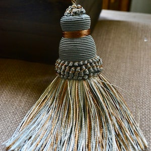 May include: A decorative tassel in shades of grey, gold, and pale blue. The tassel features a cylindrical body with a gold band and a braided top. The bottom has a cascade of matching threads. Suitable for decorative use.