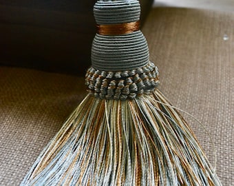 May include: Decorative tassel with a gray, gold, and light blue color scheme. The tassel has a cylindrical body with a gold band and a braided top. The bottom features a cascade of matching threads. The tassel is designed for decorative purposes.
