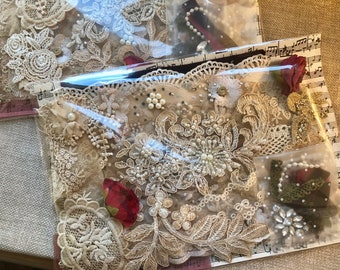 May include: Two packages of vintage lace embellishments. The lace is beige and cream-colored, with floral and pearl accents. Each package contains dried red rose buds and is wrapped in clear plastic with a musical note pattern.