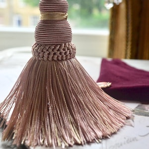 May include: A decorative tassel in shades of pink and gold, suspended from a cord. The tassel has a detailed, layered design with a braided section and a flowing fringe. The background includes a window and a burgundy fabric.