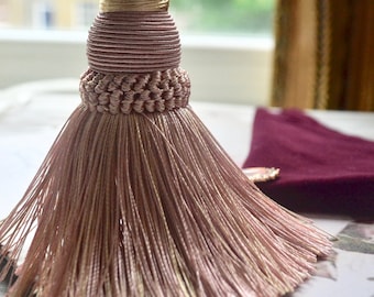 May include: A decorative tassel in shades of pink and gold, hanging from a cord. The tassel features a detailed, layered design with a braided section and a flowing fringe. The background includes a window and a burgundy fabric.