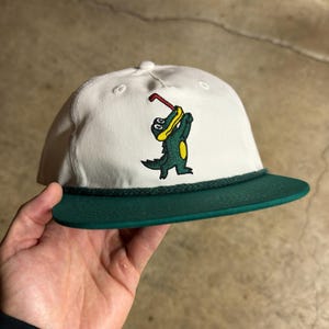 May include: A white baseball cap with a dark green brim and a matching dark green rope detail across the base of the crown. The front features an embroidered graphic of a green alligator wearing a yellow shirt and holding a red golf club.
