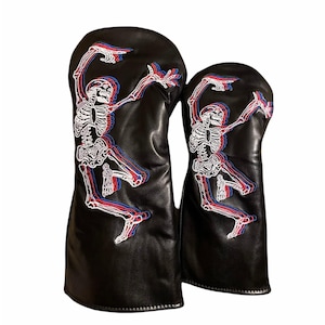May include: Two black golf club head covers with a skeleton design. The skeleton is white with red and blue outlines, giving a 3D effect. The covers are made of a smooth, leather-like material.