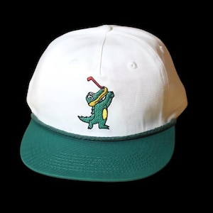 May include: White baseball cap with a green brim and an embroidered alligator holding a golf club.