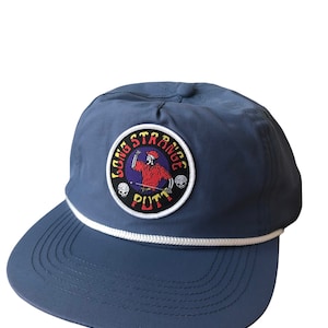 May include: Navy blue cap with a white rope detail and a flat brim. The cap features a circular patch with the text "LONG STRANGE PUTT" and a graphic of a skeleton golfer. The design includes red, white, and black colors.