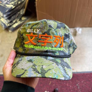 May include: A camouflage print baseball cap featuring the white text BILLY, large orange Japanese characters, and the green text BLUEGRASS MUSIC embroidered on the front. The cap includes a black braided cord trim across the base of the crown.
