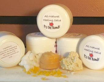May include: Four white jars of all-natural healing salve with red and white labels that say "All-Natural Healing Salve It's the Bomb!"  The jars are arranged in a pyramid shape.  The jars are sitting on a white surface with a small amount of the salve in each jar.