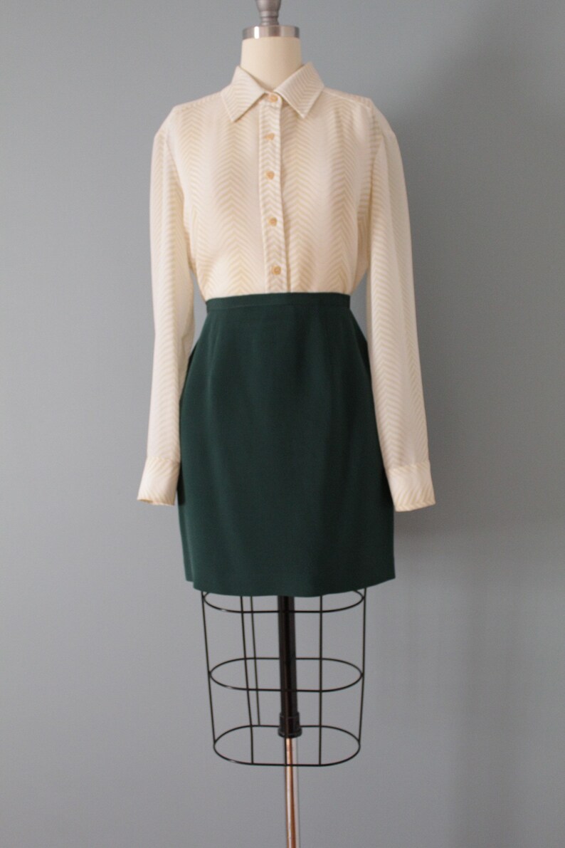 May include: A white button-down shirt with a chevron pattern and a dark green skirt. The shirt has long sleeves and a collared neckline. The skirt is knee-length and has a straight silhouette.