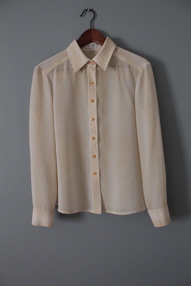 May include: A white long-sleeved button-down shirt with a yellow chevron pattern. The shirt has a collar and is hanging on a wooden hanger.