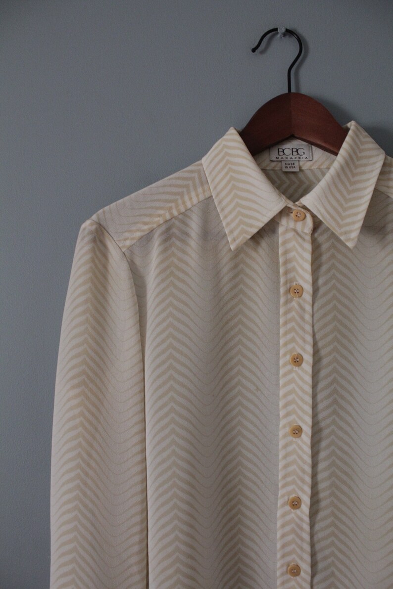 May include: A white long-sleeved button-down shirt with a chevron pattern. The shirt has a collar and is made of a silky fabric. The brand label reads "BCBG Max Azria" and "Made in USA".