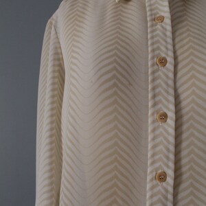 May include: A long-sleeved, cream-colored button-down shirt with a chevron pattern. The shirt has a classic collar and a button-up front.