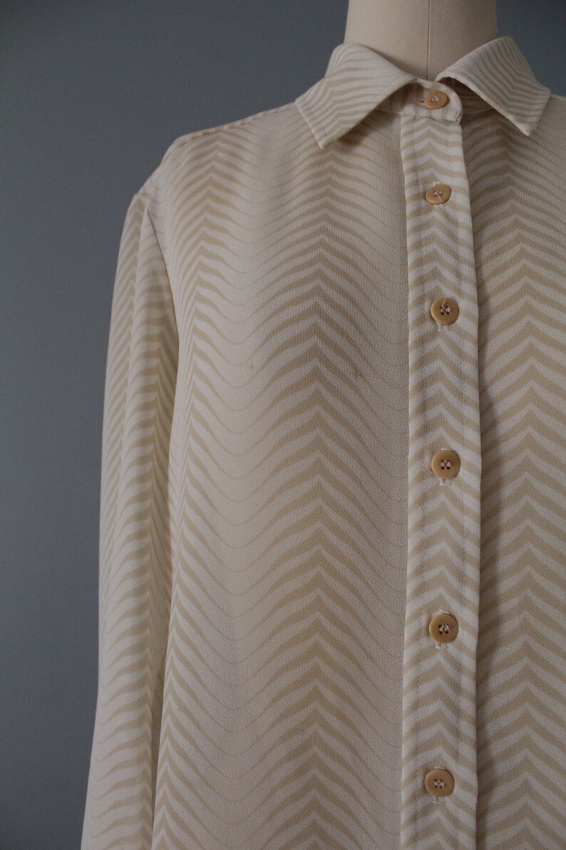 May include: A long-sleeved, cream-colored button-down shirt with a chevron pattern. The shirt has a classic collar and a button-up front.