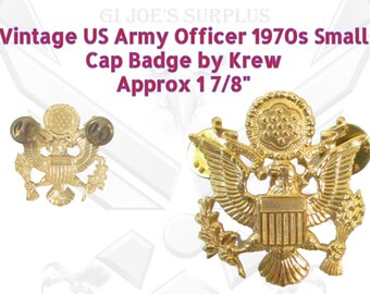 May include: A gold-colored US Army Officer cap badge from the 1970s. The badge features an eagle with a shield and a star, and is approximately 1 7/8 inches in size. The badge is made by Krew.