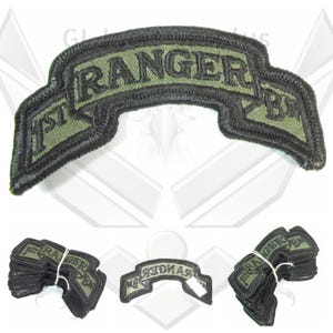 May include: A black and olive green embroidered patch with the text "1st RANGER BN". The patch has a curved banner shape with a black border. Several stacks of the same patch are also visible.