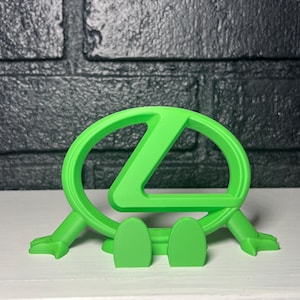 May include: A bright lime green plastic phone stand featuring the stylized Lexus logo as the main frame. The design includes two small feet at the front and two extended arms at the sides, creating a stable base for holding a mobile device.