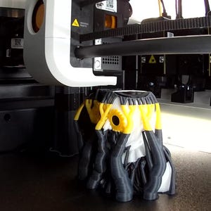 May include: A 3D printer in operation, featuring a white print head positioned above a partially completed object. The object is composed of black, white, and yellow filament, displaying a complex, multi-layered structure on the print bed.