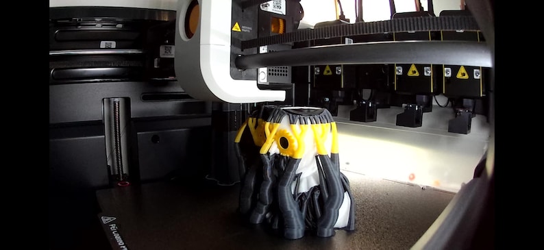 May include: A 3D printer in operation, featuring a white print head positioned above a partially completed object. The object is composed of black, white, and yellow filament, displaying a complex, multi-layered structure on the print bed.