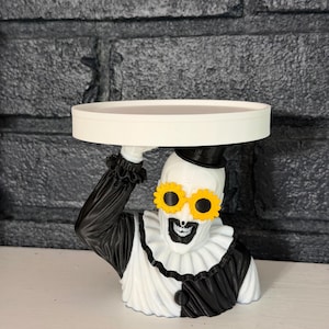 May include: A decorative stand featuring a sculpted clown figure wearing a black top hat, a black and white ruffled collar, and yellow flower-shaped sunglasses. The clown figure holds a flat, circular white tray above its head with one hand.