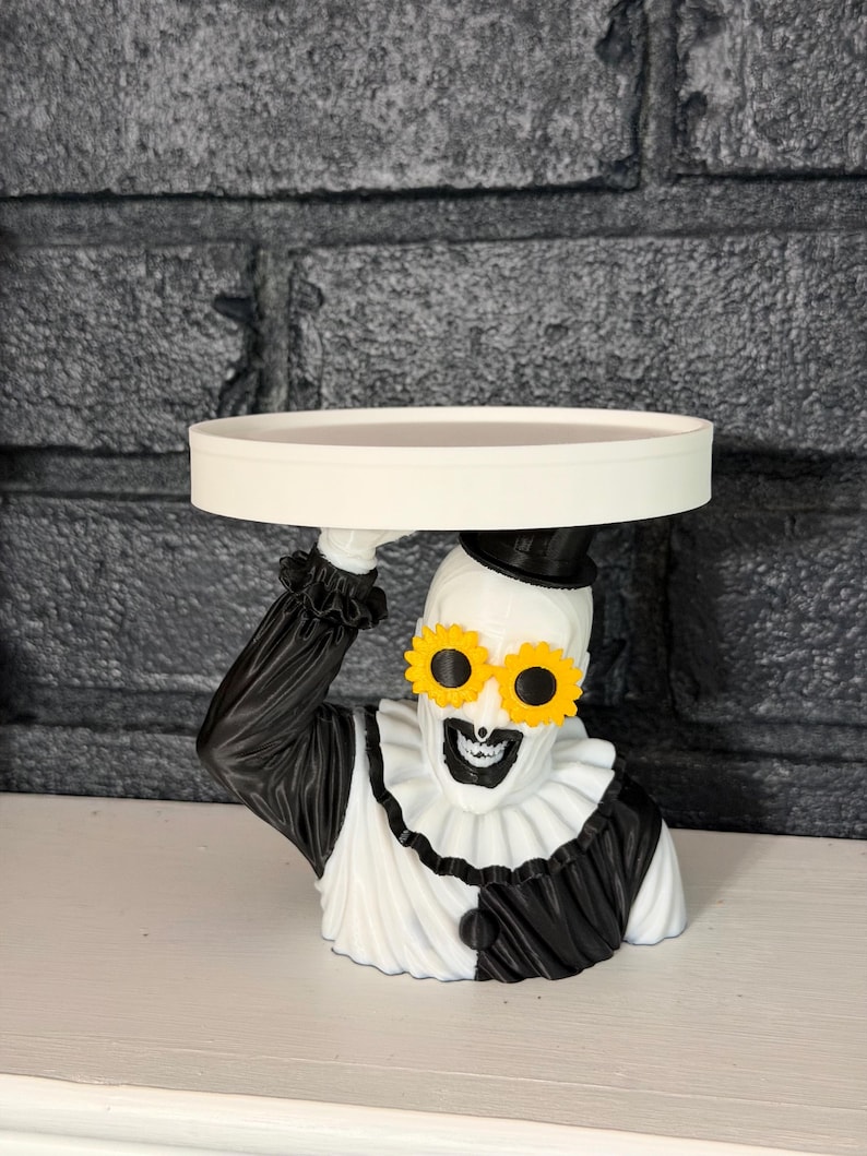 May include: A decorative stand featuring a sculpted clown figure wearing a black top hat, a black and white ruffled collar, and yellow flower-shaped sunglasses. The clown figure holds a flat, circular white tray above its head with one hand.