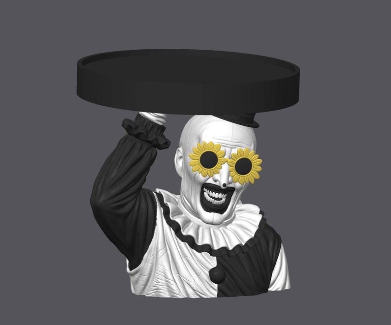 May include: A black and white clown bust featuring a wide grin and yellow sunflower-shaped sunglasses. The clown wears a ruffled collar and holds a flat, circular black tray above its head with one raised arm, showcasing a detailed monochromatic design.
